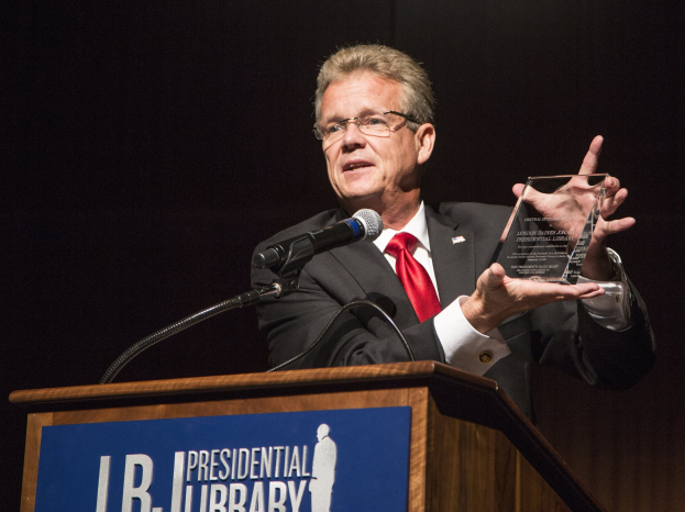 Ein Mann im Anzug und mit Krawatte, der eine Auszeichnung an einem Podium mit Mikrofon und Tafel hält, vor einem dunklen Hintergrund bei einer Bibliothek-Veranstaltung.