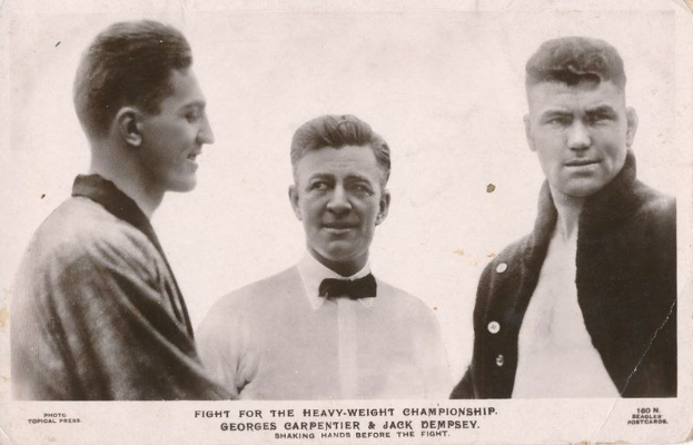 Drei Männer stehen nebeneinander auf einem Schwarz-Weiß-Foto, wobei der Mann in der Mitte ein weißes Hemd und eine Krawatte trägt, flankiert von zwei Männern in Anzügen, und der Text 'Kampf um die Schwergewichtmeisterschaft' unten.