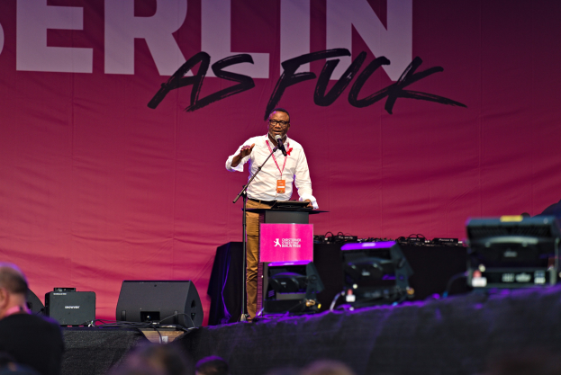 Ein Mann in einem weißen Hemd und braunen Hosen steht auf einer Bühne und hält ein Mikrofon in der Hand, mit einem pinken "Berlin as Fuck"-Schild hinter ihm und einer Menge vor ihm.