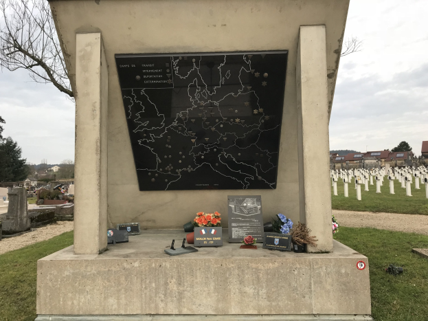 Ein Friedhofdenkmal mit einer Karte, umgeben von Blumen und Gegenständen, das das Grab eines deutschen Weltkriegs-Soldaten markiert, mit Gräbern, Bäumen, Gebäuden und einem blauen Himmel im Hintergrund.