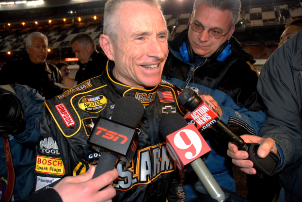 Ein Mann in einer schwarzen Jacke spricht mit Reportern vor einem hell erleuchteten Stadion voller Zuschauer, einige halten Mikrofone und einer eine Kamera.