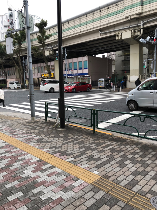 Eine Stadtstraße mit Autos, Fußgängern, die an einer Ampel über die Straße gehen, einem Fußweg, Geländer, Pfosten, Verkehrszeichen, einer Brücke, Bäumen, Gebäuden mit Fenstern und einem klaren blauen Himmel.