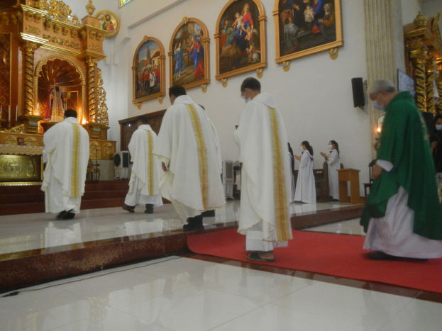 Eine Gruppe von Priestern geht einen roten Teppich in einer Kirche entlang, mit Menschen auf dem Boden und auf der rechten Seite, und Foto Rahmen an der Wand im Hintergrund.