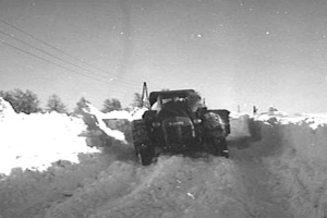 Ein Schwarz-Weiß-Foto eines Traktors, der eine verschneite Straße entlangfährt, mit Bäumen und Drähten im Hintergrund.