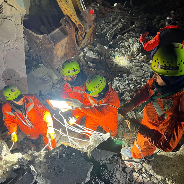 Gruppe von Menschen in orangefarbenen Uniformen und grünen Helmen bei der Arbeit an Schutt mit einem Kran im Hintergrund während einer Rettungsoperation.