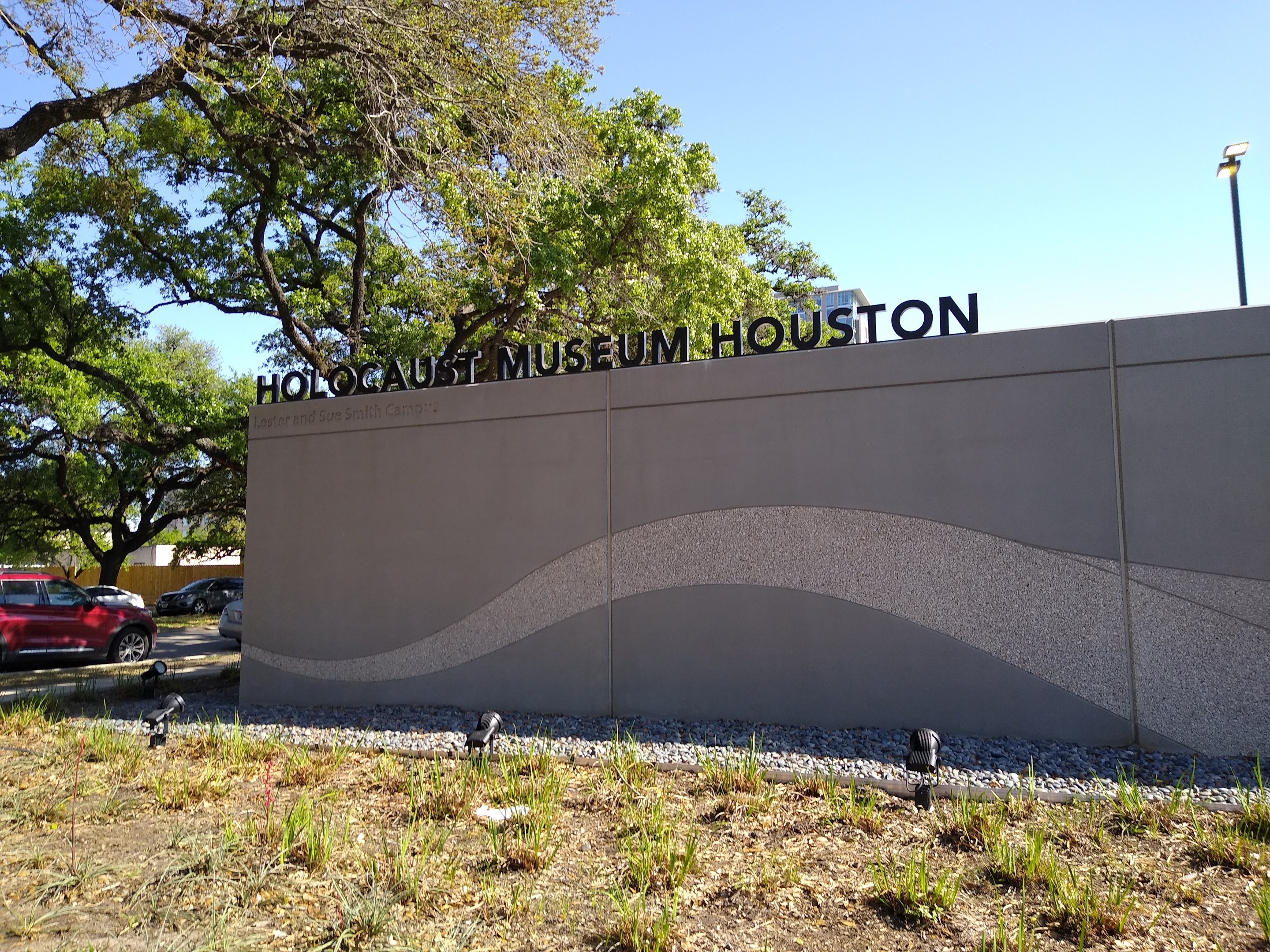 Außenansicht des Houston Holocaust Museums mit eingravierter Textwand, verstreuten Steinen, Grünfläche, Bäumen, vorbeifahrenden Fahrzeugen, einem Straßenschild und einem bewölkten Himmel.
