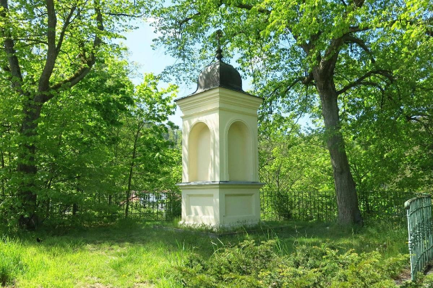 Kleines weißes Holocaust-Denkmal auf einem grasbewachsenen Friedhof, umgeben von einem Zaun und Bäumen, unter einem klaren blauen Himmel in Vilnius, Litauen.