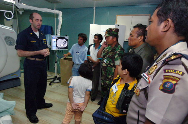 Eine Gruppe von Menschen steht um ein Krankenhauszimmer herum, ein Mann hält in der Mitte ein Mikrofon, ein Bett mit einer Decke darauf links, verschiedene medizinische Geräte, ein Röntgengerät, eine Tafel mit Text, eine Tür, eine Wand und eine Decke mit Deckenleuchten im Hintergrund.