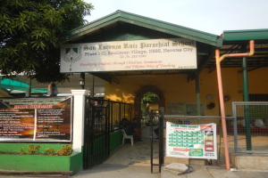 Der Eingang der San Lorenzo Ruiz Pfarrschule hat ein Schild mit Text, einen Metallzaun, Menschen auf Stühlen unter einem Dach, Pfosten, ein Hinweisschild, Pflanzen, einen Baum und einen bewölkten Himmel.