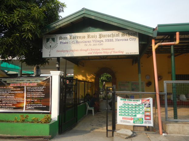 Der Eingang der San Lorenzo Ruiz Pfarrschule hat ein Schild mit Text, einen Metallzaun, Menschen auf Stühlen unter einem Dach, Pfosten, ein Hinweisschild, Pflanzen, einen Baum und einen bewölkten Himmel.