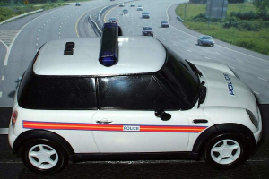 Ein weißes Polizeiauto steht im Vordergrund mit vielen Autos auf der Straße dahinter, umgeben von Gras und Bäumen auf beiden Seiten.