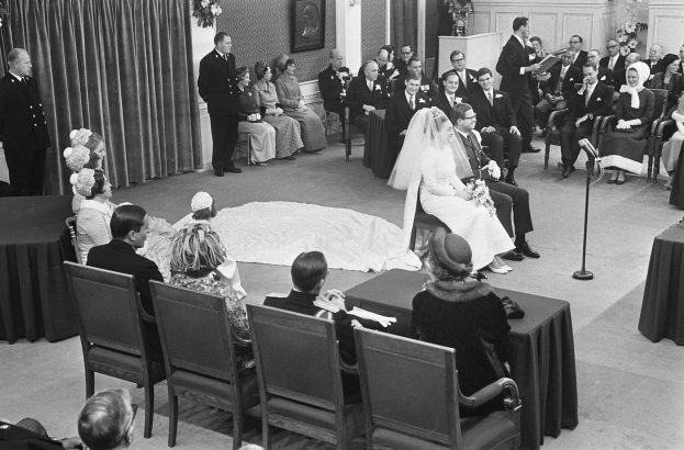 Schwarzes und weißes Foto einer Hochzeitszeremonie mit sitzenden und stehenden Menschen, einem Mann mit Mikrofon, einer Frau mit Blumenstrauß, einem Podium mit Mikrofon, Blumengestecken, einem Vorhang, einem gerahmten Foto an der Wand und einer Tür im Hintergrund.