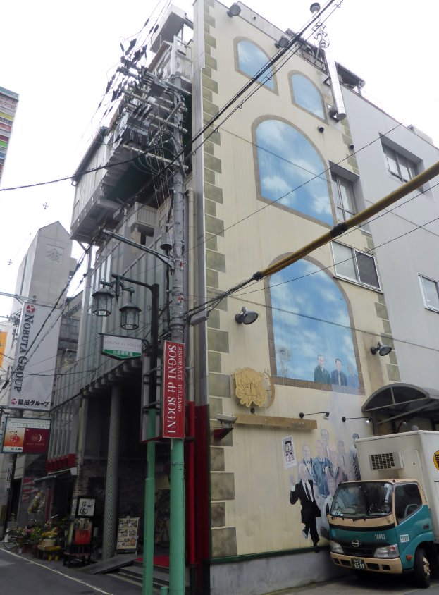 Ein Lastwagen parkt an der Seite einer Straße neben einem hohen Gebäude mit einer Wandmalerei, mit Strommasten, Kabeln, Straßenlaternen, Namensschildern, Kraftfahrzeugen, Topfpflanzen und einem klaren blauen Himmel im Hintergrund.