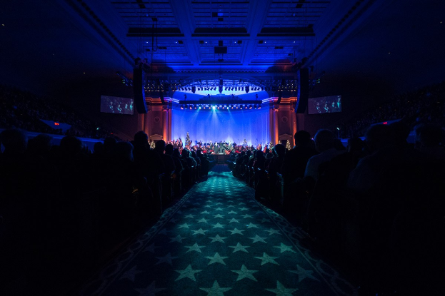 Ein großer Saal mit Menschen in Reihen vor einer Bühne, auf der Musiker spielen, unter einer Decke mit Lichtern und Fernsehern, was auf eine Konzerthalle für die Nationale Convention 2019 hinweist.