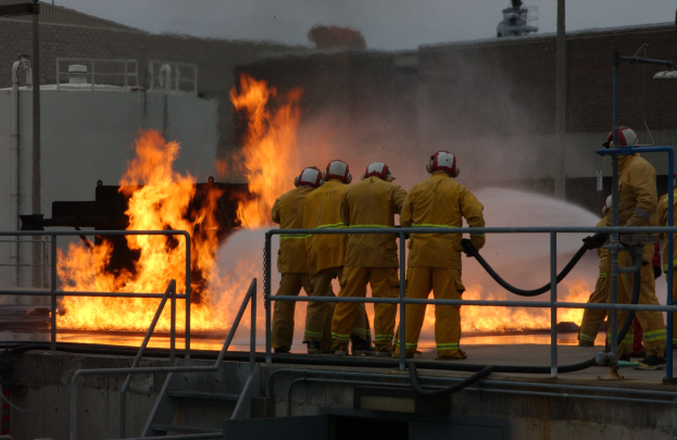 Firefighters in helmets stand on a rooftop holding hoses, with railings, steps, a building, and sky in the background.