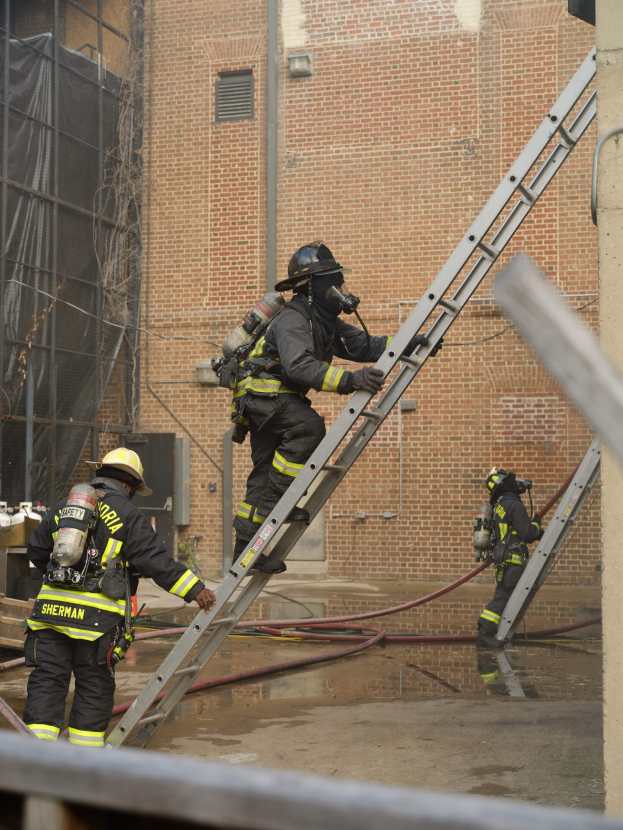 Feuerwehrleute in Helmen und Luftzylindern klettern an einer Leiter vor einem Backsteingebäude mit Rohren auf dem Boden und einer Metallstange unten, während im Hintergrund ein weiteres Gebäude mit Fenstern und einem Netz zu sehen ist.
