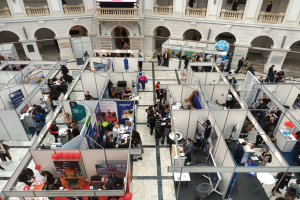 Eine große Gruppe von Menschen versammelt sich in einem Raum mit Ständen, Bannern, Tischen, Stühlen, Ballons und einem Gebäude mit Bögen, Säulen, Geländern und einem Bildschirm im Hintergrund.