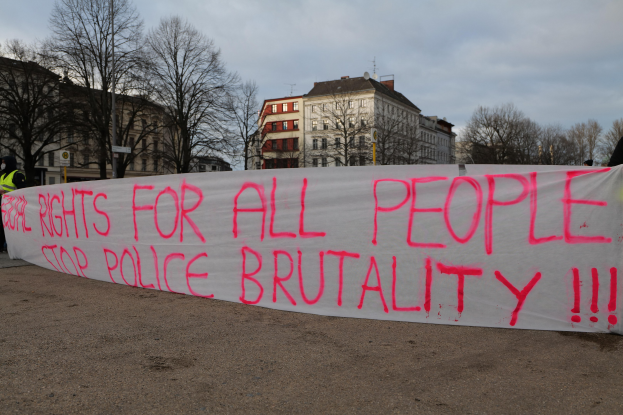 Eine Gruppe von Menschen hält ein Transparent mit der Aufschrift 'Rechte für alle Menschen Stoppt Polizeigewalt' vor einem Straßenschild, einer Litfaßsäule, Bäumen, Gebäuden mit Fenstern und einem bewölkten Himmel.