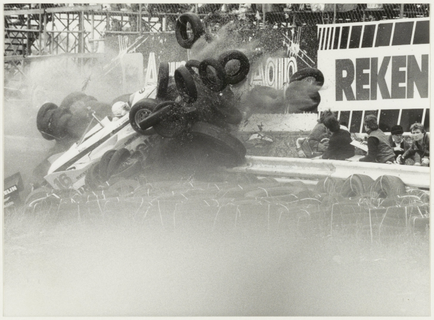 Schwarzes und weißes Foto eines verunglückten Rennwagens, umgeben von Reifen, mit Zuschauern auf der rechten Seite und einer Tafel mit Text im Hintergrund.