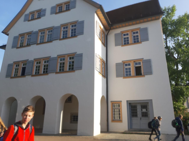 Eine Gruppe von Menschen, die vor einem großen weißen Gebäude mit Fenstern und Türen entlanggeht, umgeben von Bäumen unter einem klaren blauen Himmel, einige tragen Taschen.