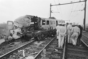 Eine Schwarz-Weiß-Szene einer Zugkollision mit einer Gruppe von Menschen in Helmen drumherum, mit Strommasten, Bäumen, Gebäuden und einem klaren Himmel im Hintergrund.