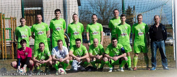 Eine Gruppe von Männern in grünen T-Shirts steht und hockt auf einem Fußballfeld mit einem Ball davor, ein Torpfosten rechts und ein Hintergrund aus Bäumen, Gebäuden, Pfählen, Fahrzeugen und dem Himmel, mit Text in der linken unteren Ecke, der die Zahlen von 1 bis 72 auflistet.