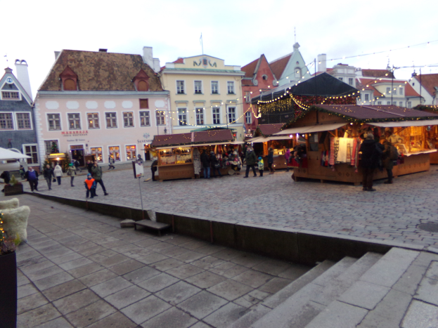 Ein geschäftiger Weihnachtsmarkt in Tallinn, Estland, mit Menschen umstände, die mit Lichtern, festlichen Gegenständen und Topfpflanzen geschmückt sind, vor Gebäuden mit Fenstern unter einem bewölkten Himmel, der über Stufen erreichbar ist.