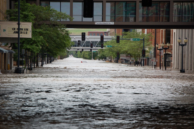 Eine überflutete städtische Straße mit Wasser, das die Straße, die Infrastruktur und die umliegenden Gebäude bedeckt, mit einer Brücke im Hintergrund.