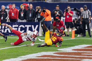 Ein Football-Spieler taucht in die Endzone für ein Touchdown, mit einer Gruppe von Menschen am Boden, einige halten Kameras, und eine Fahne mit Text im Vordergrund und Geländern, Pfosten und andere Gegenstände im Hintergrund.
