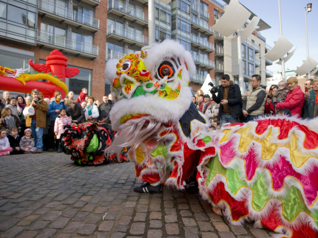 Ein lebendiges chinesisches Neujahrsfest in Amsterdam mit einer Löwendance-Vorführung und einer Menge Zuschauern, einige halten Kameras, vor einem Hintergrund aus Gebäuden, Laternenmästen und einem klaren blauen Himmel.