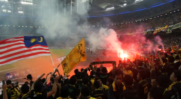 Eine große Menschenmenge in einem Stadion mit Bengalos, Fahnen und einer Kamera im Hintergrund, mit Rauch und Schildern, auf denen "Malaysia vs Malaysia Live-Streaming" steht.