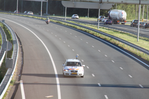 Ein Auto fährt auf einer Autobahn mit einem Polizeiwagen am Rande, anderen Fahrzeugen, Gelöndern, Pfosten, Planken und Bäumen im Hintergrund.