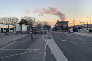 Eine Stadtstraße mit Absperrbaken, einem Bus, Laternen, Bäumen, Gebäuden, Kränen und Rauch, der aus einem fernen Gebäude aufsteigt.