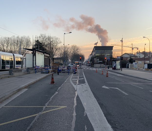 Eine Stadtstraße mit Absperrbaken, einem Bus, Laternen, Bäumen, Gebäuden, Kränen und Rauch, der aus einem fernen Gebäude aufsteigt.