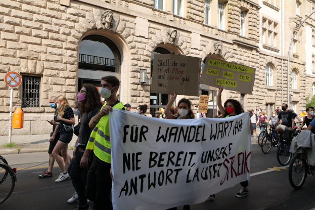 Eine Gruppe von Menschen marschiert auf einer Straße, einige halten Protestschilder und andere fahren Fahrräder, im Hintergrund ist ein historisches Gebäude mit Rundbogenfenstern, Säulen und Skulpturen zu sehen.
