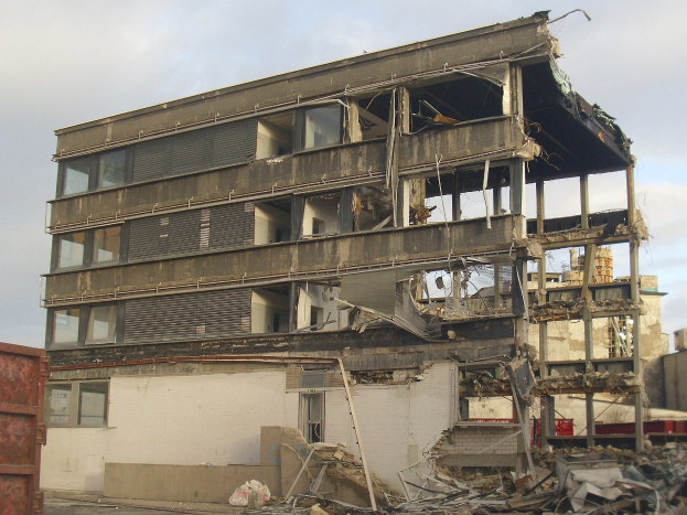 Ein teilweise abgerissenes Gebäude mit zerbrochenen Fenstern und Schutt in der Mitte einer Straße, mit einem Container links und einem bewölkten Himmel im Hintergrund.