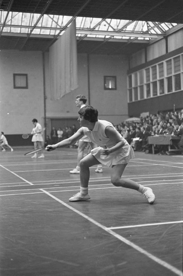 Schwarzes und weißes Foto von zwei Frauen, die Badminton auf einem Platz spielen, jede hält einen Schläger in der Hand, mit Zuschauern rechts und einer Wand mit Fenstern im Hintergrund.