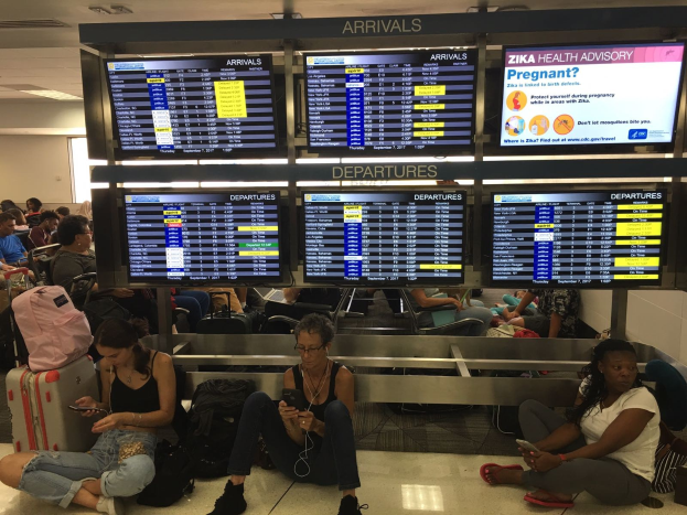 Menschen sitzen auf dem Flughafenboden, einige halten Handys, mit Taschen drumherum und Bildschirmen im Hintergrund, die Zika-Virus-Informationen anzeigen.