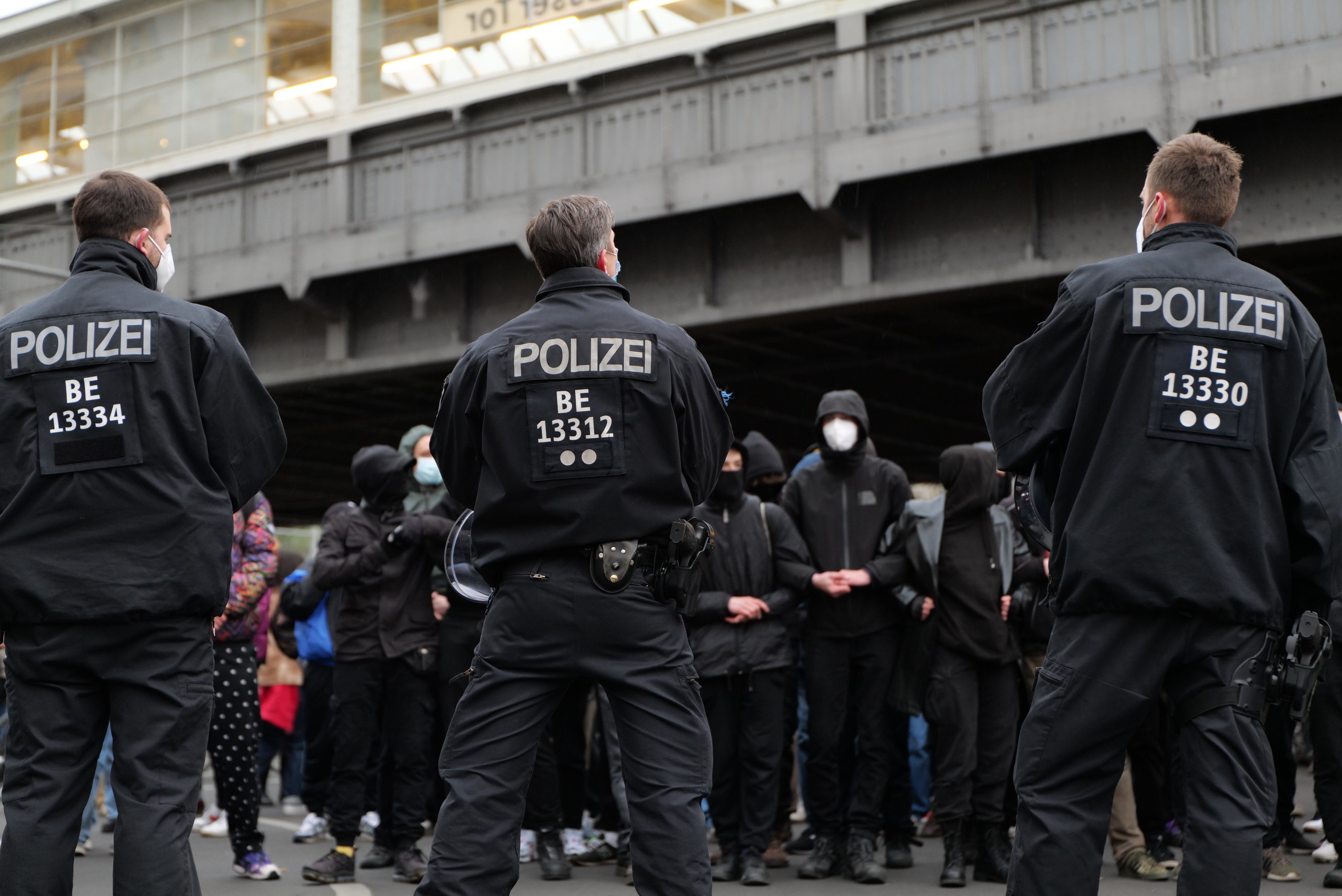 Eine Gruppe von Polizisten in Uniform steht vor einer Menge von Menschen in schwarzen Uniformen und Masken, mit einer Stadtbrücke und einem Gebäude im Hintergrund, während einer Demonstration.