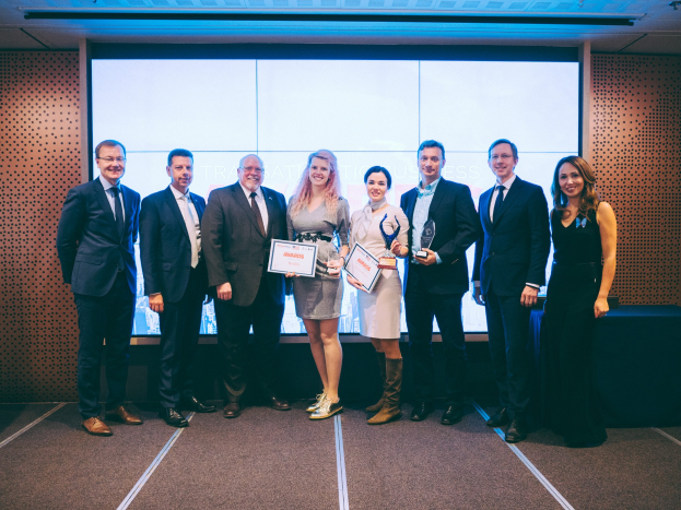 Eine Gruppe strahlender Menschen mit Urkunden, die vor einem Bildschirm stehen, mit einem Tisch und einem wandmontierten Objekt im Hintergrund, als Gewinner der International Business Awards 2018 ausgezeichnet.