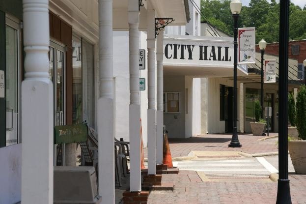 Ein Rathausgebäude mit umliegender Infrastruktur, darunter Straßenmasten, Lichter, Namensschilder, Fahrzeuge, Verkehrskegel, Stühle, Topfpflanzen, Bäume und einen bewölkten Himmel.