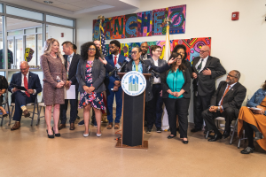 Gruppe von Menschen vor einem Podium mit einem Mikrofon und einer Tafel mit der Aufschrift "Sancho City Councilman Joins City Council", mit sitzenden und stehenden Zuschauern, Flaggen, einem Feuerlöscher und Deckenleuchten im Hintergrund.