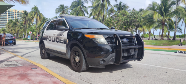 Polizeiauto auf der Straßenseite mit Menschen, Fahrrädern, Gras, Bäumen, Gebäuden und einem klaren blauen Himmel im Hintergrund; Vorderrad sichtbar.