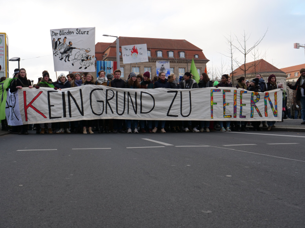 Demonstranten auf einer Straße mit einem Banner mit der Aufschrift "Kein Grund zu Feiern" gegen deutsche Sparmaßnahmen, mit Gebäuden, Bäumen und einem klaren Himmel im Hintergrund.
