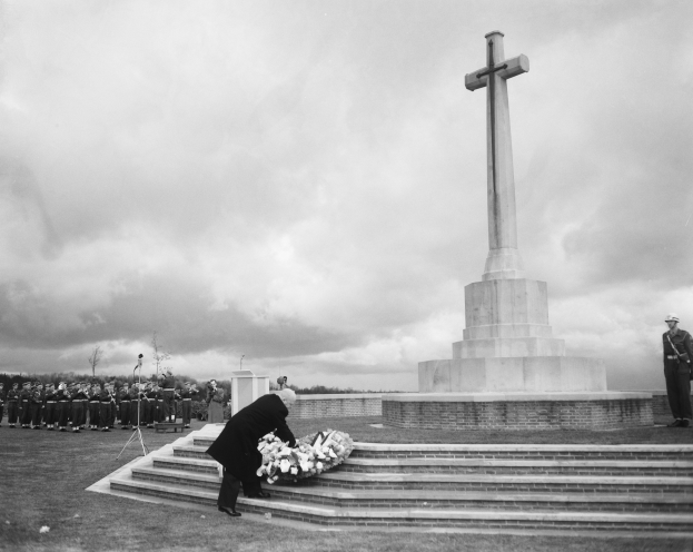 Ein Mann legt einen Kranz auf die Stufen eines Weltkriegsdenkmals, das von einer Gruppe Menschen umgeben ist, mit Bäumen und einem bewölkten Himmel im Hintergrund, dargestellt in Schwarz-Weiß.