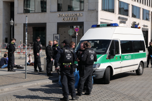 Eine Gruppe von Polizeibeamten steht vor einem Starbucks-Café, rechts daneben ein Van geparkt, links Menschen und im Hintergrund ein Gebäude mit Fenstern, einer Schautafel, einem Laternenmast und einem Zaun.