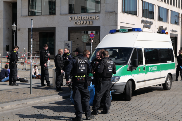 Eine Gruppe von Polizeibeamten steht vor einem Starbucks-Café, rechts daneben ein Van geparkt, links Menschen und im Hintergrund ein Gebäude mit Fenstern, einer Schautafel, einem Laternenmast und einem Zaun.