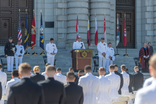 Gruppe von Männern in weißen Uniformen vor einem Gebäude mit Säulen und Türen, mit einem Podium und Mikrofon in der Mitte, Flaggen im Hintergrund und Stühlen und Treppen, die zum Gebäude führen.