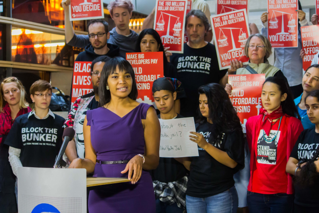 Black Lives Matter-Aktivistin spricht vor Publikum mit Mikrofon, das Plakate haltende Menge, vor Lichtern und anderen Gegenständen.