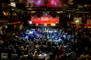 Eine große Menschenmenge sitzt auf Stühlen vor einer Bühne, die von Scheinwerfern beleuchtet und von Geländern umgeben ist. Auf der Bühne befinden sich Bildschirme und Banner mit Text, und in der linken unteren Ecke befindet sich ein Wasserzeichen, was eine Atmosphäre der Aufregung und Vorfreude für ein Online-Marketing-Rockstars-Event schafft.